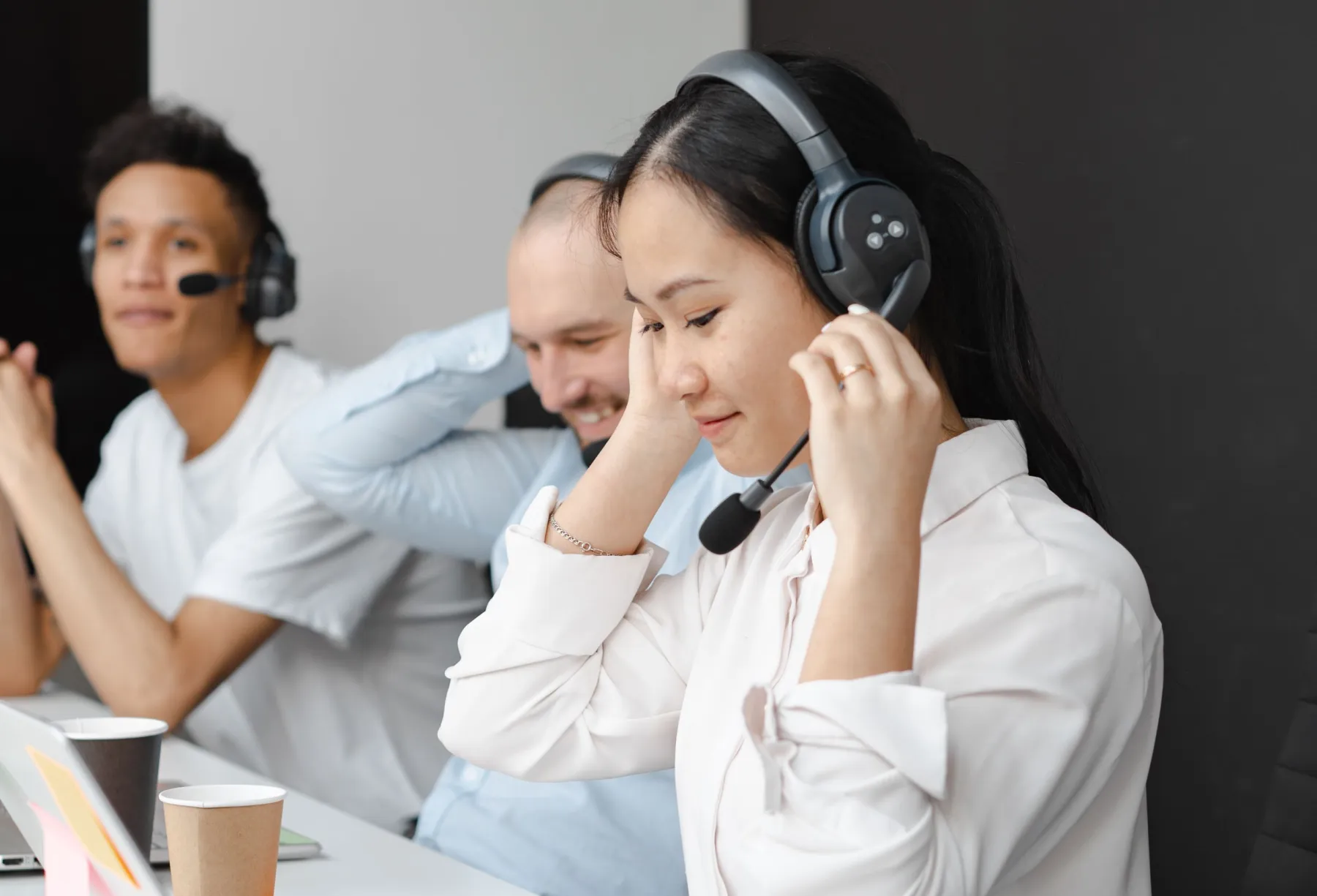 Three contact center team members wearing headsets at a shared desk, one adjusting her headset while a colleague smiles